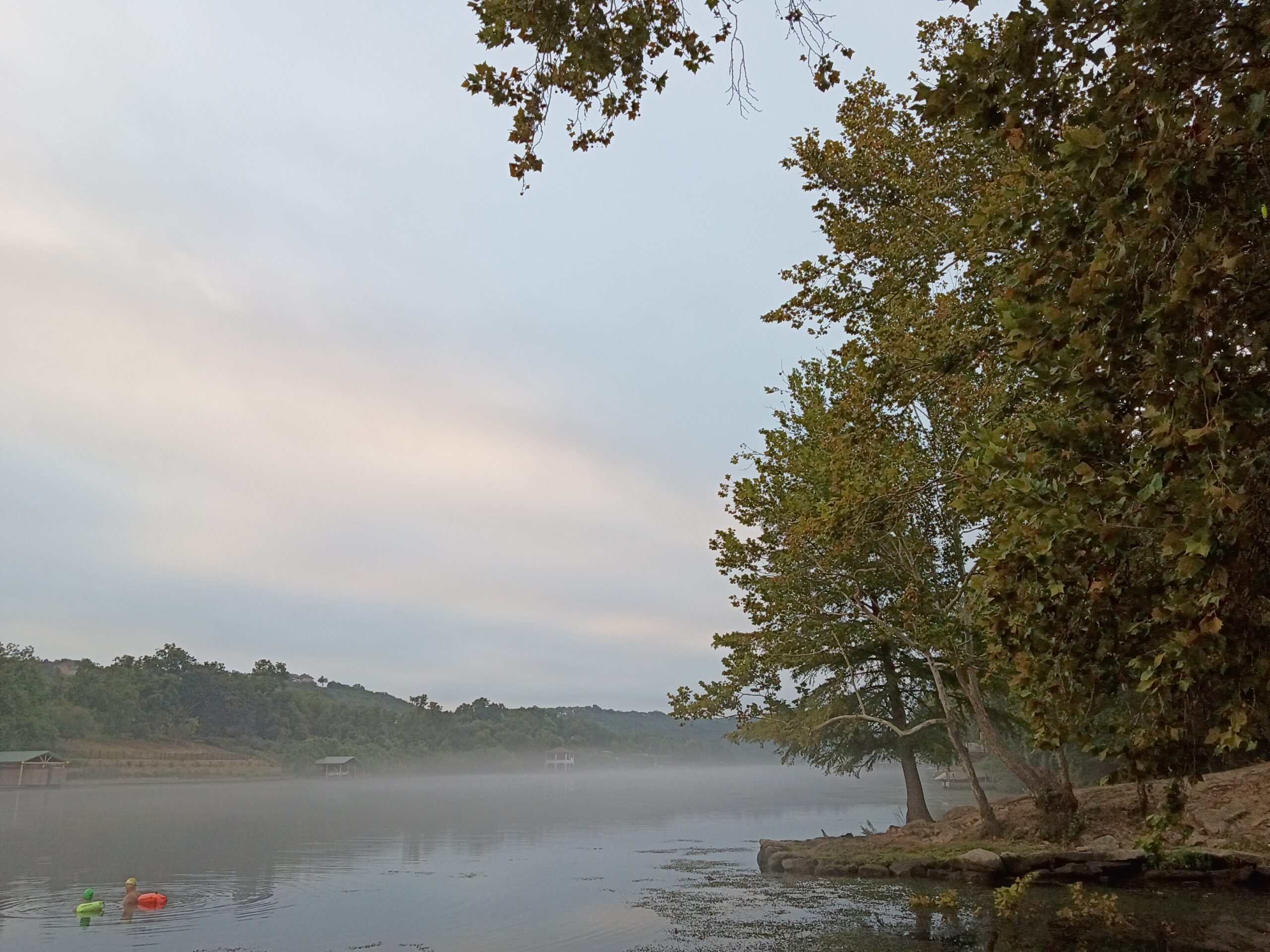 Wide view across the water at Lakeside Park, with two swimmers at the far right holding onto a buoy and the wooded shoreline in the distance.