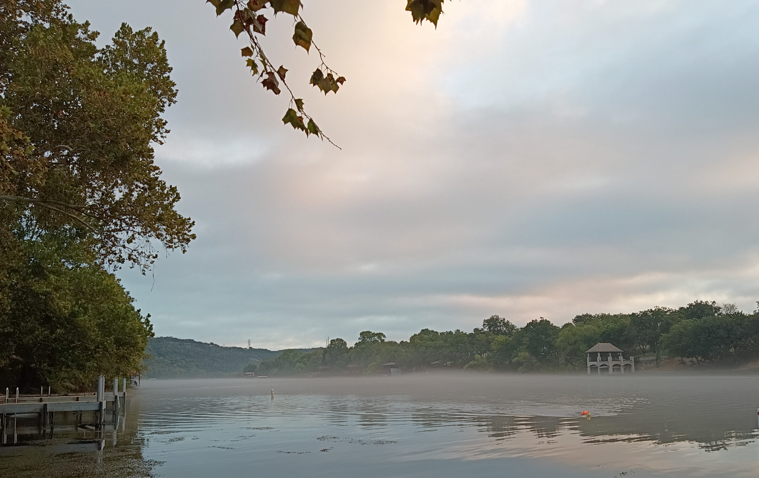 Mist floating just above Lake Austin on a quiet early morning, with tree-lined shores and soft pink light appearing through the clouds.